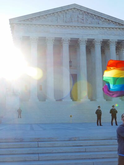 A man waves a rainbow flag in front of the steps of the Supreme Court.