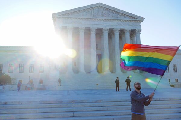 A man waves a rainbow flag in front of the steps of the Supreme Court.
