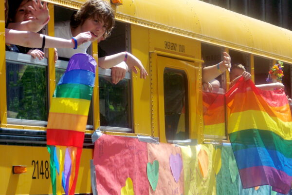 Children wave rainbow flags from a yellow school-bus.