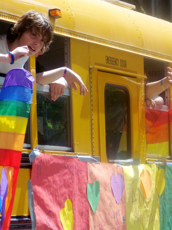 Children wave rainbow flags from a yellow school-bus.