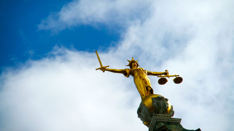 A golden statue holding the scales of justice in one hand and a sword in the other against a blue sky backdrop.