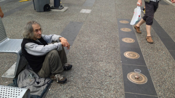 A man panhandles on the side of a public sidewalk as people walk by.
