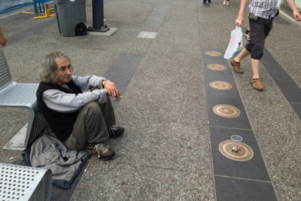 A man panhandles on the side of a public sidewalk as people walk by.