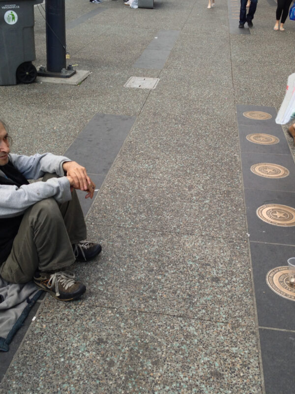 A man panhandles on the side of a public sidewalk as people walk by.
