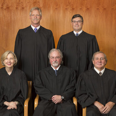 The Alaska Supreme Court justices (four men and one one woman) sit on a bench, all wearing black judicial robes.