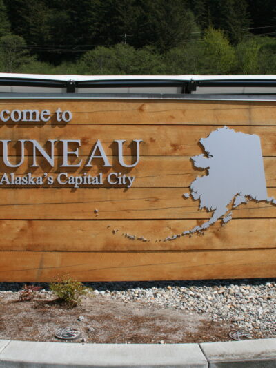 A wooden sign says "Welcome to Juneau - Alaska's Capital City" next to a map of Alaska