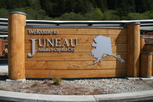 A wooden sign says "Welcome to Juneau - Alaska's Capital City" next to a map of Alaska