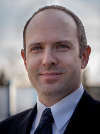 Executive Director Joshua Decker stands against a blue sky backdrop wearing an ACLU lapel pin.