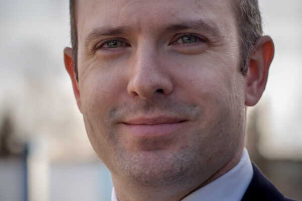 Executive Director Joshua Decker stands against a blue sky backdrop wearing an ACLU lapel pin.
