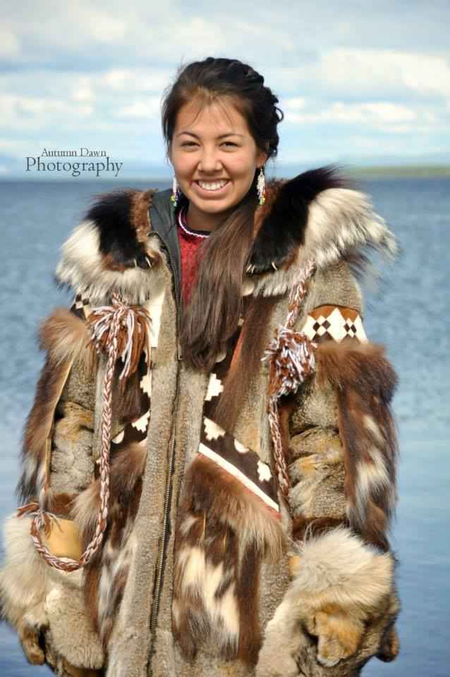 A young woman dressed in traditional Alaska Native clothing and furs stands against a body of water.