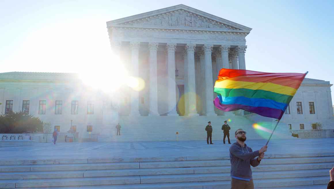 A man waves a rainbow flag in front of the steps of the Supreme Court.