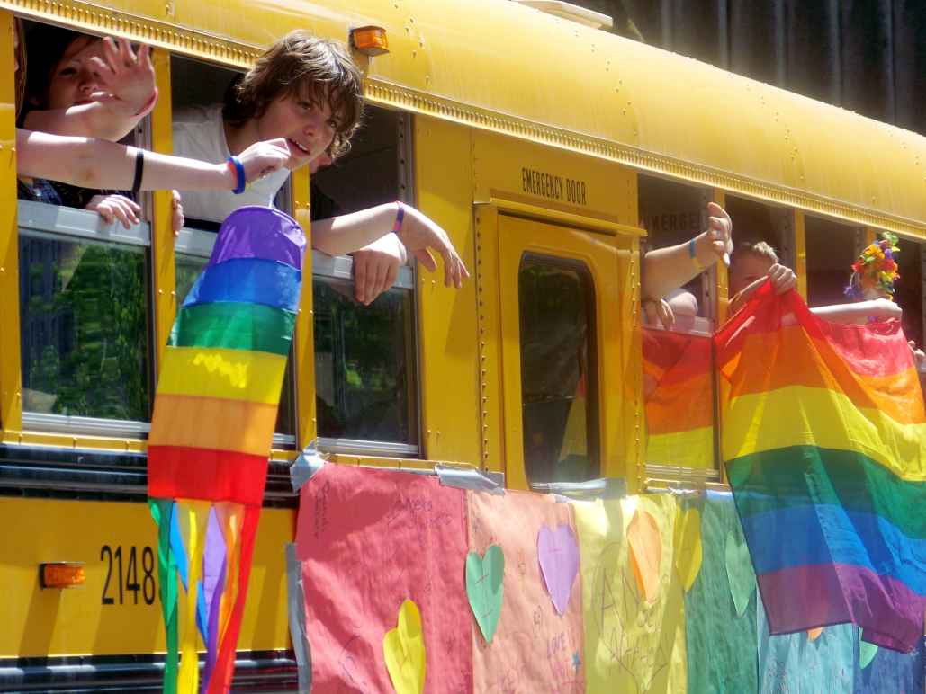 Children wave rainbow flags from a yellow school-bus.