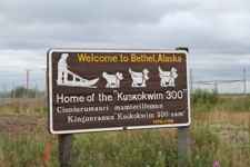 A brown sign standing in a field says "Welcome to Bethel, Alaska" above white pictures of Iditarod sled-dogs.