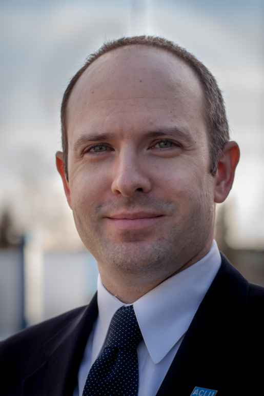 Executive Director Joshua Decker stands against a blue sky backdrop wearing an ACLU lapel pin.