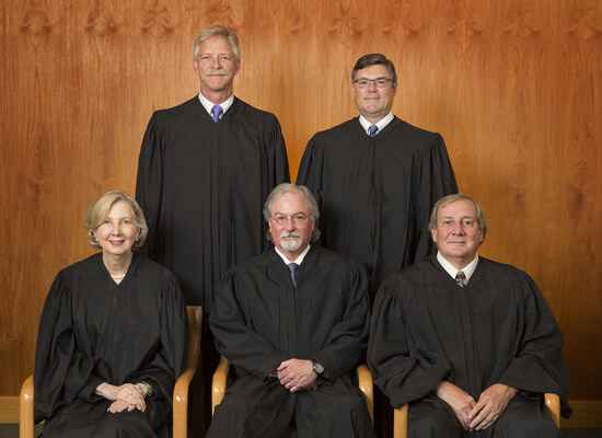 The Alaska Supreme Court justices (four men and one one woman) sit on a bench, all wearing black judicial robes.