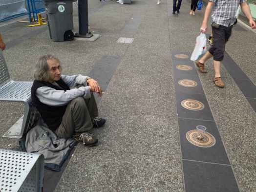 A man panhandles on the side of a public sidewalk as people walk by.