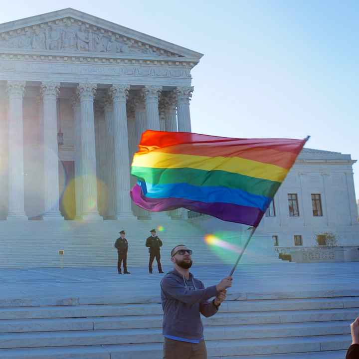 A man waves a rainbow flag in front of the steps of the Supreme Court.