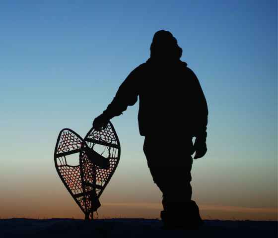 A man is pictured from behind holding a pair of snowshoes against a sunset backdrop.